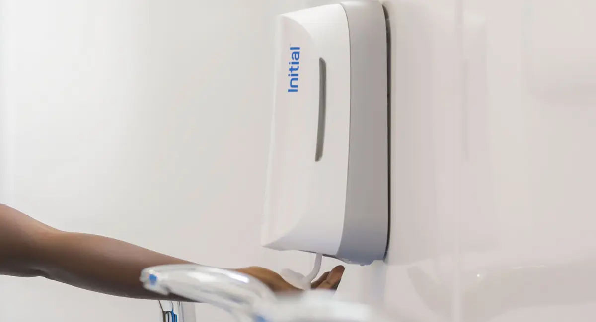 A hand receiving soap from a wall-mounted Initial soap dispenser next to a sink faucet.
